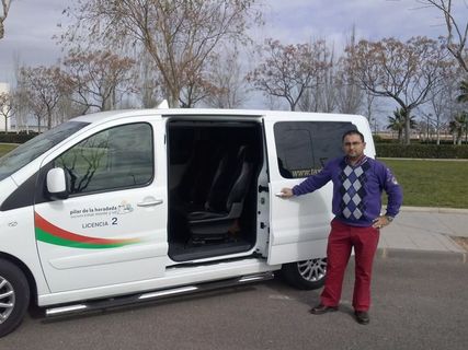 A person stands next to an open white passenger van in a park, wearing a patterned sweater and red trousers.
