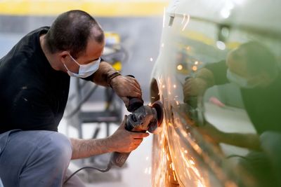 Person mit Mundschutz benutzt einen Winkelschleifer, um am Radkasten eines Autos zu arbeiten, wobei helle Funken entstehen.