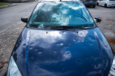 Dunkelblauer Wagen mit zersplitterter Windschutzscheibe und zahlreichen Hagelschäden auf der Motorhaube.
