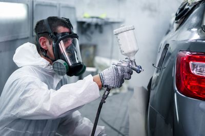 Person,die eine Vollgesichtsmaske und einen Schutzanzug trägt, sprüht in einer Werkstatt Farbe auf die Rückseite eines Autos.