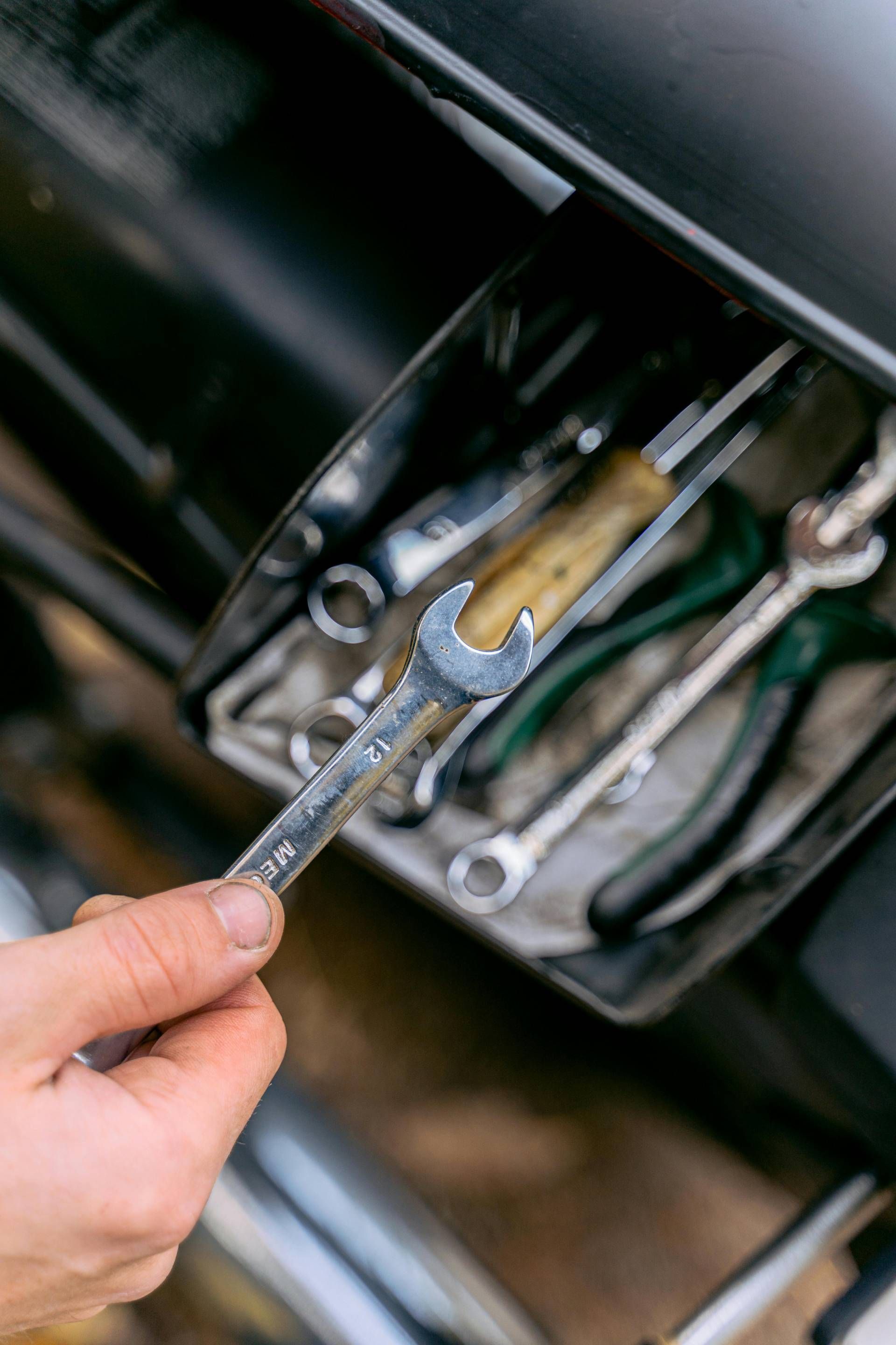 Hand hält einen Metallschlüssel vor einem offenen Werkzeugkasten, der verschiedene, sortierte Handwerkzeuge enthält.