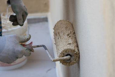 Une main gantée utilise un rouleau à peinture pour recouvrir un mur extérieur beige.