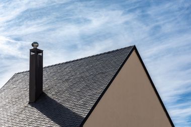 Toit de maison en tuiles sombres avec une cheminée se détachant sur un ciel bleu.