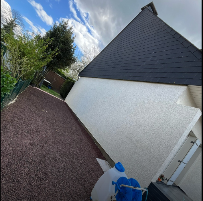Vue inclinée de l'extérieur d'une maison avec un chemin de gravier, un mur blanc, un toit foncé et un conteneur bleu au premier plan.