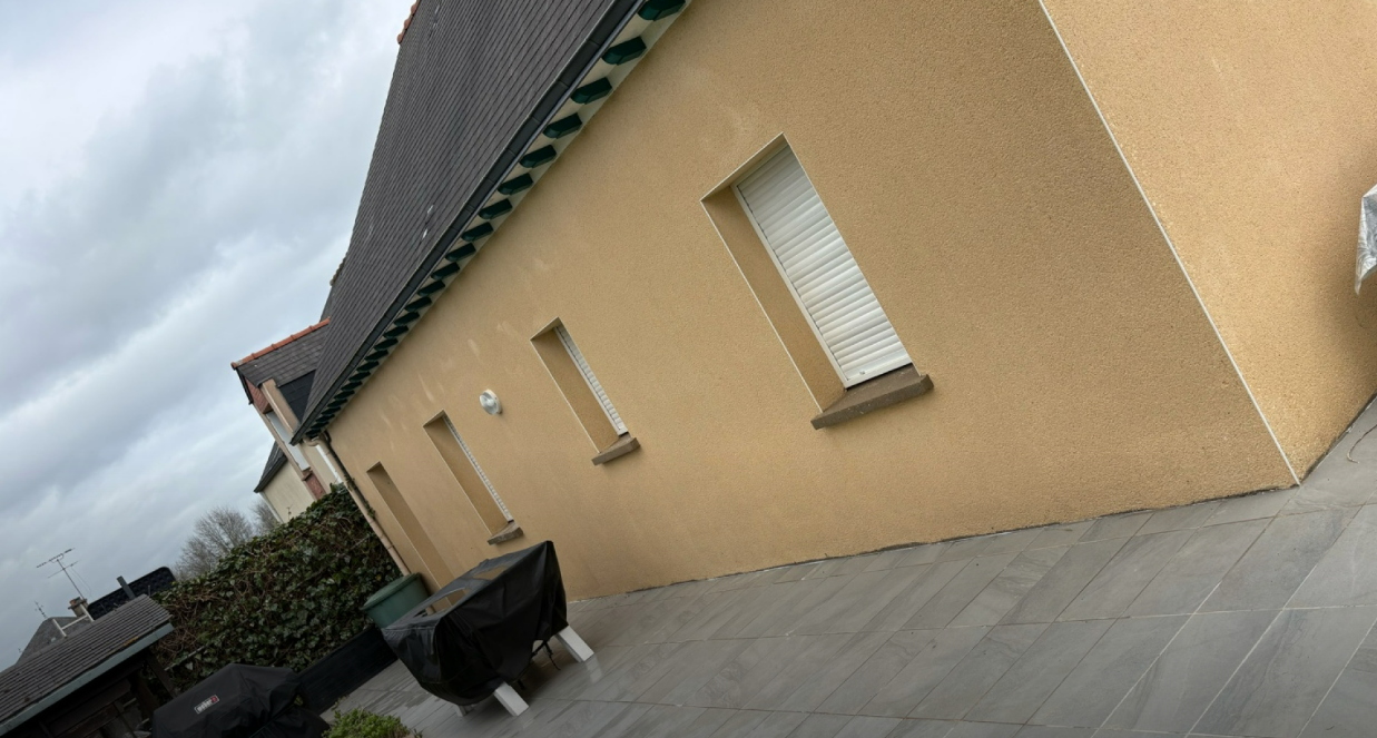 Vue inclinée d'une maison beige au toit gris, avec de petites fenêtres, une terrasse pavée et une poubelle sous un ciel nuageux.