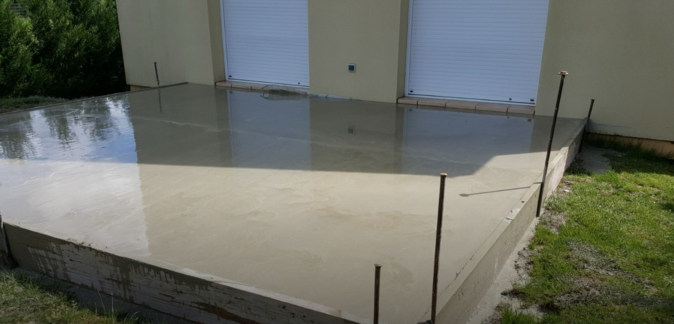 Dalle de béton inondée à côté de portes de garage blanches et de pelouse, l'eau stagnante reflétant la lumière du soleil.