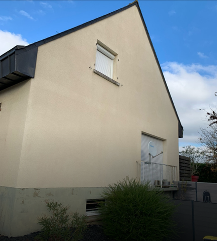 Maison crème à deux étages avec un toit pentu, de petites fenêtres et une cour avant clôturée sous un ciel bleu