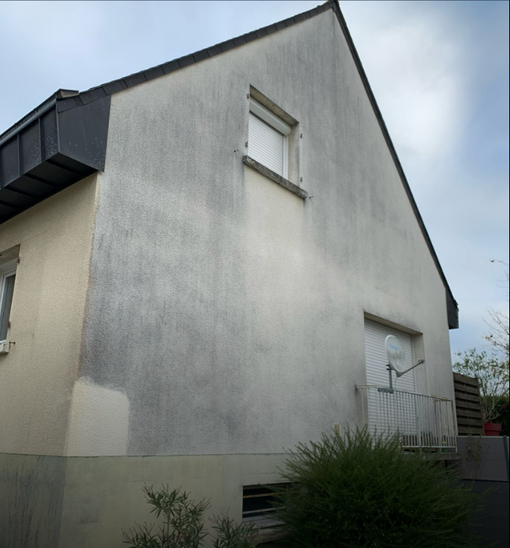 Maison à l'extérieur en stuc gris, avec de petites fenêtres et un toit en pente, sous un ciel nuageux.