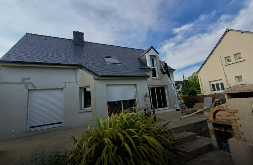 Maison avec toit en ardoise, puits de lumière et escalier de terrasse donnant sur un jardin surélevé sous un ciel nuageux.