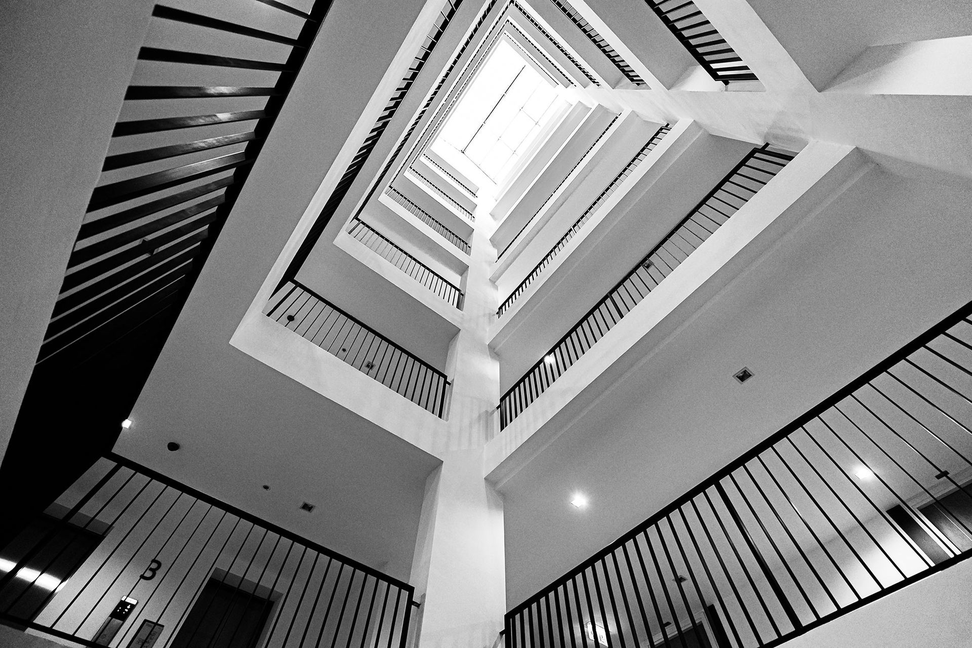 Schwarz-Weiß-Fotografie mit Blick in ein offenes Treppenhaus. Mehrere Stockwerke mit schwarzen Geländern.