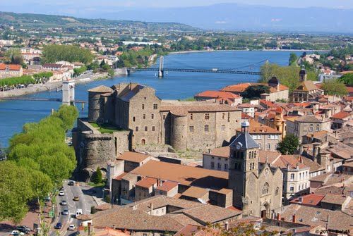 Forteresse historique en pierre surplombant une rivière et une ville aux toits rouges dans une vallée ensoleillée