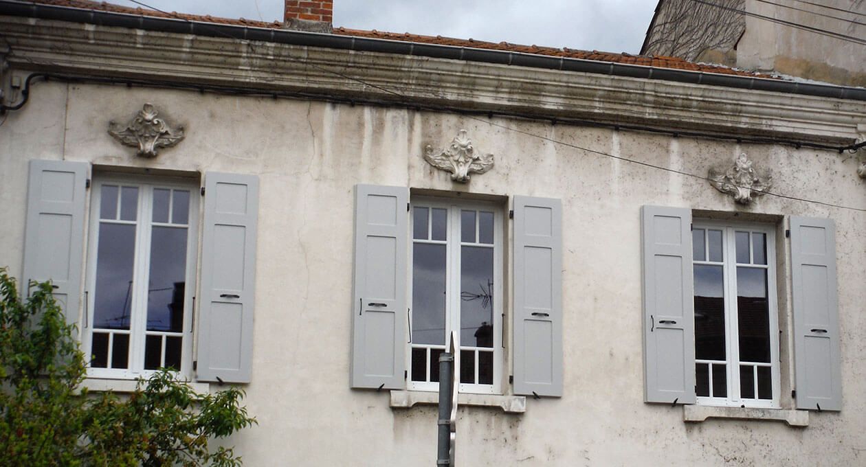 Trois volets blancs fermés sur la façade d'un bâtiment en stuc patiné par le temps, avec un bord de toit en tuiles.