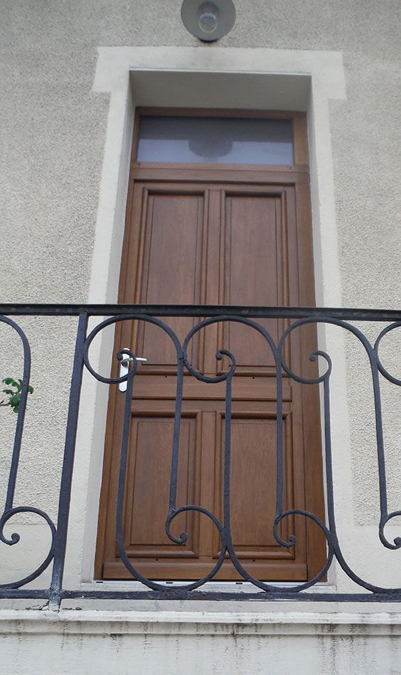 Porte en bois avec imposte vitrée et balustrade de balcon en fer forgé noir sur un mur extérieur gris