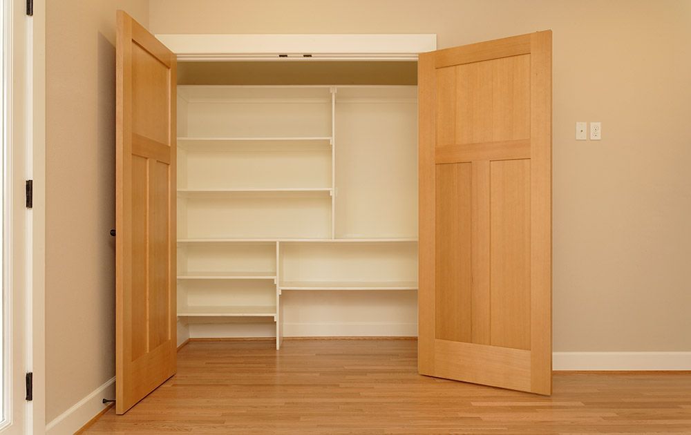 Ouvrez les doubles portes en bois qui révèlent un placard intégré vide avec des étagères blanches et un sol en parquet.