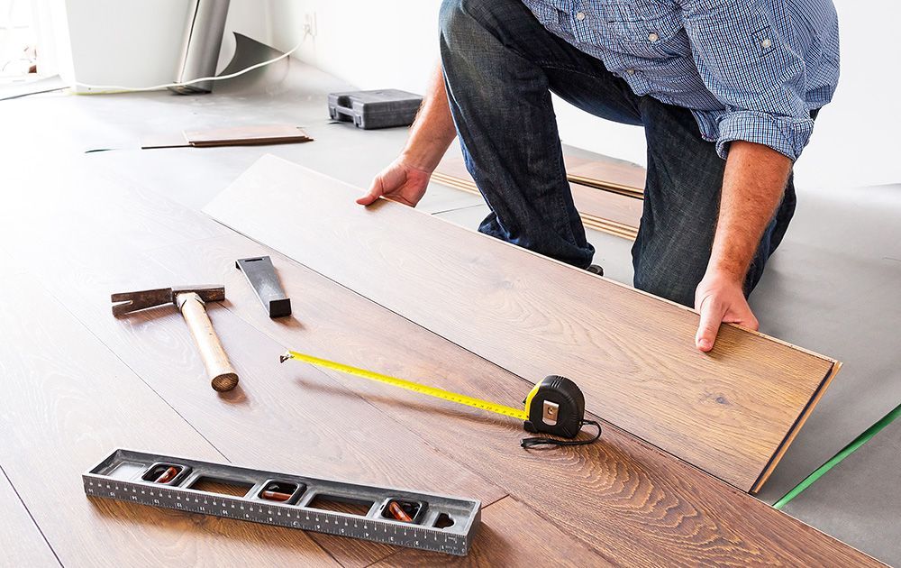 Une personne installe des lames de parquet à l'aide d'un marteau, d'un mètre ruban et d'un niveau sur un sol blanc.