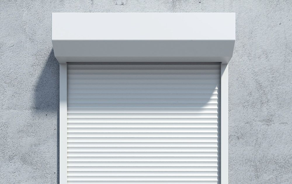Volet roulant blanc sur un mur gris avec un petit auvent au-dessus.