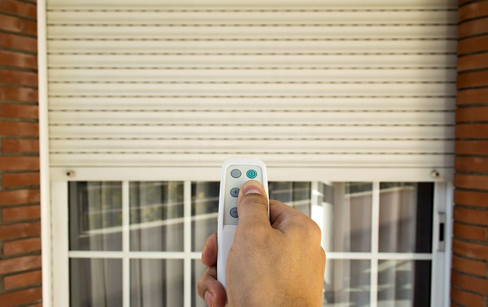Main tenant une télécommande, pointée vers un volet roulant d'un bâtiment en briques