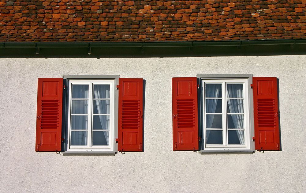 Mur blanc avec deux fenêtres, chacune encadrée de volets rouges, sous un toit de tuiles.