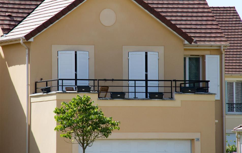 Extérieur d'une maison beige avec balustrade de balcon noire, volets blancs et un arbre devant.