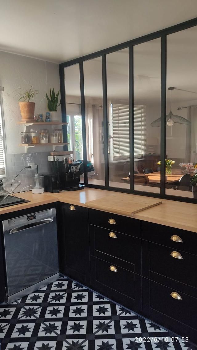 Cuisine noire avec robinetterie dorée, sol en carrelage à damier et cloison vitrée donnant sur un salon.