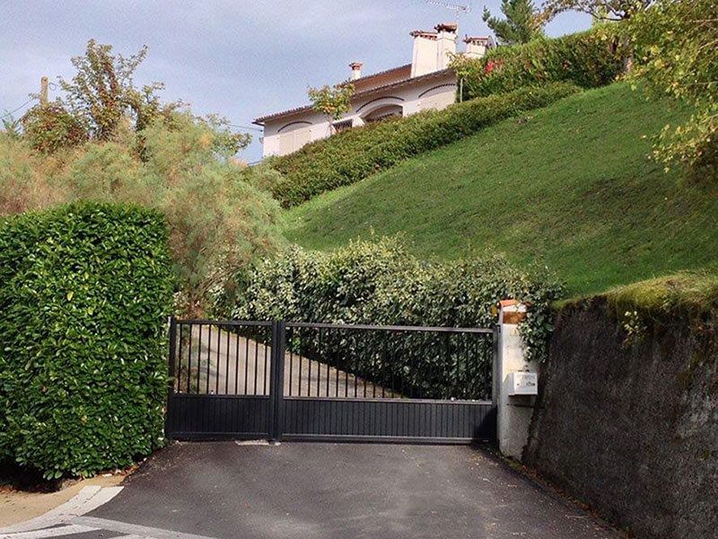 Un portail noir fermé au bout d'une allée, au pied d'une colline herbeuse et d'une maison.