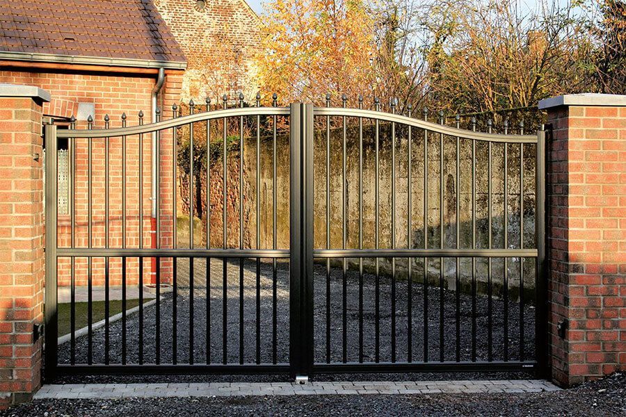 Portail d'allée en métal noir entre des piliers de brique devant une maison et des arbres d'automne