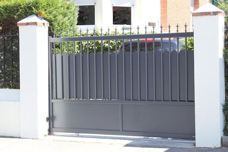 Portail d'allée en métal gris entre des piliers blancs et des poteaux en brique devant une maison