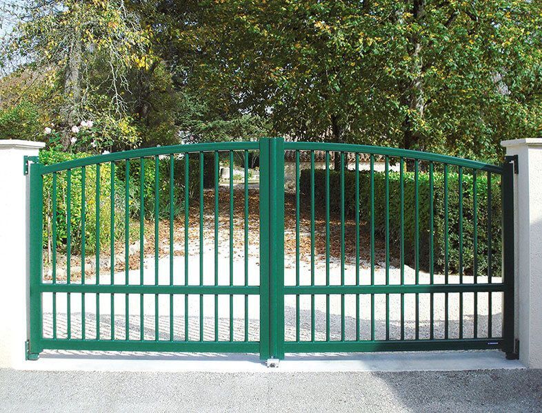 Portail d'allée en métal vert entre des piliers blancs, avec des arbres et un chemin derrière.