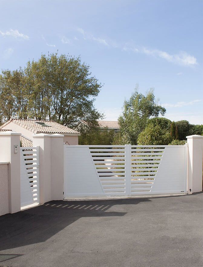 Portail et mur blancs devant une maison, avec des arbres et un ciel bleu clair en arrière-plan.