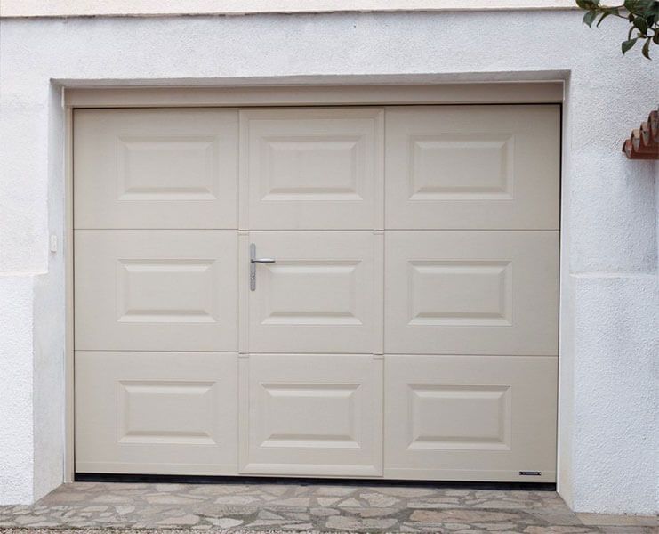 Porte de garage fermée à panneaux beiges encastrée dans un mur en stuc blanc, avec une petite poignée et une allée en pierre en dessous.