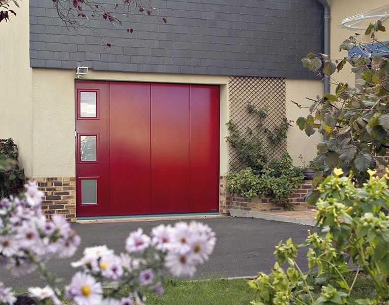Porte de garage rouge sur une maison, encadrée par des fleurs et de la verdure du jardin.