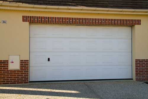 Porte de garage blanche sur une maison beige avec des bordures en briques rouges et une allée en gravier.