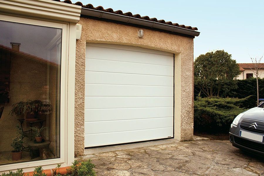 Porte de garage blanche sur une maison beige avec une voiture garée à côté d'une allée en pierre.