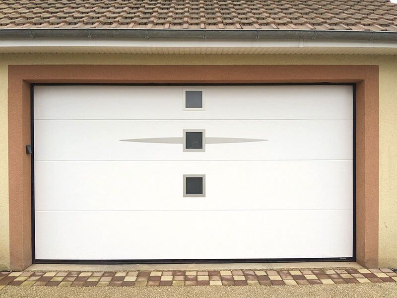 Porte de garage blanche avec petites fenêtres carrées et poignée horizontale argentée sur un mur en stuc beige.
