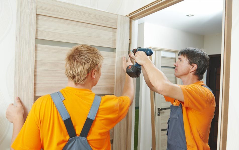 Deux ouvriers en chemises orange installent un cadre de porte en bois à l'aide d'une perceuse électrique