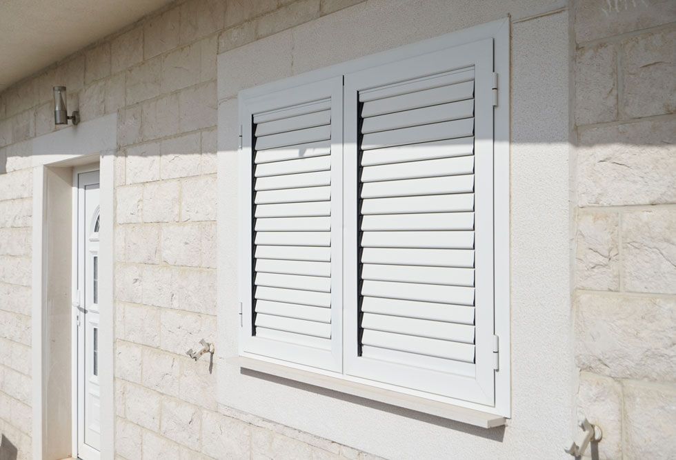 Volets persiennes blancs sur un mur extérieur en pierre beige, à côté d'une porte.