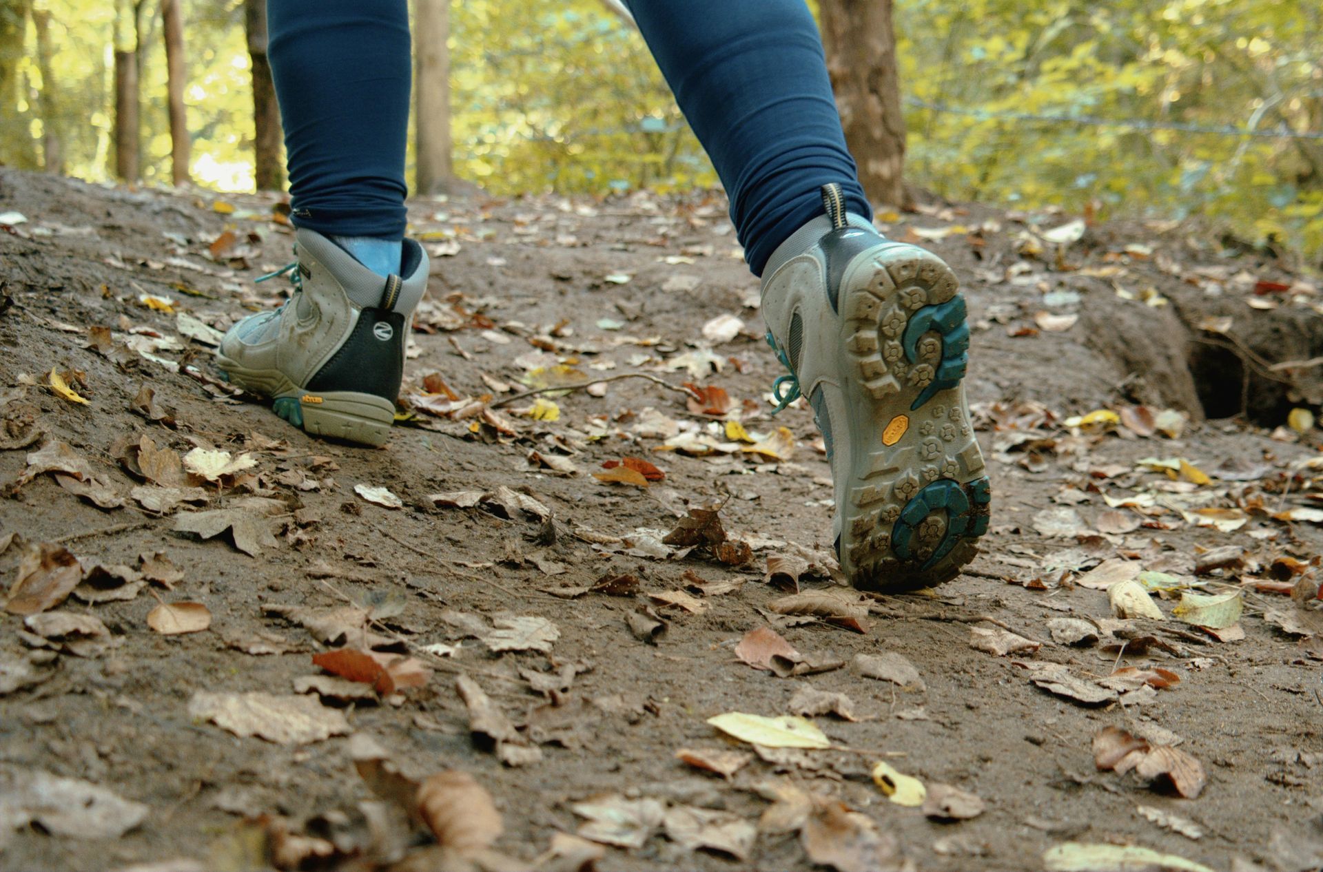 Person walking on a dirt trail, wearing hiking boots, surrounded by fallen leaves in a forest.