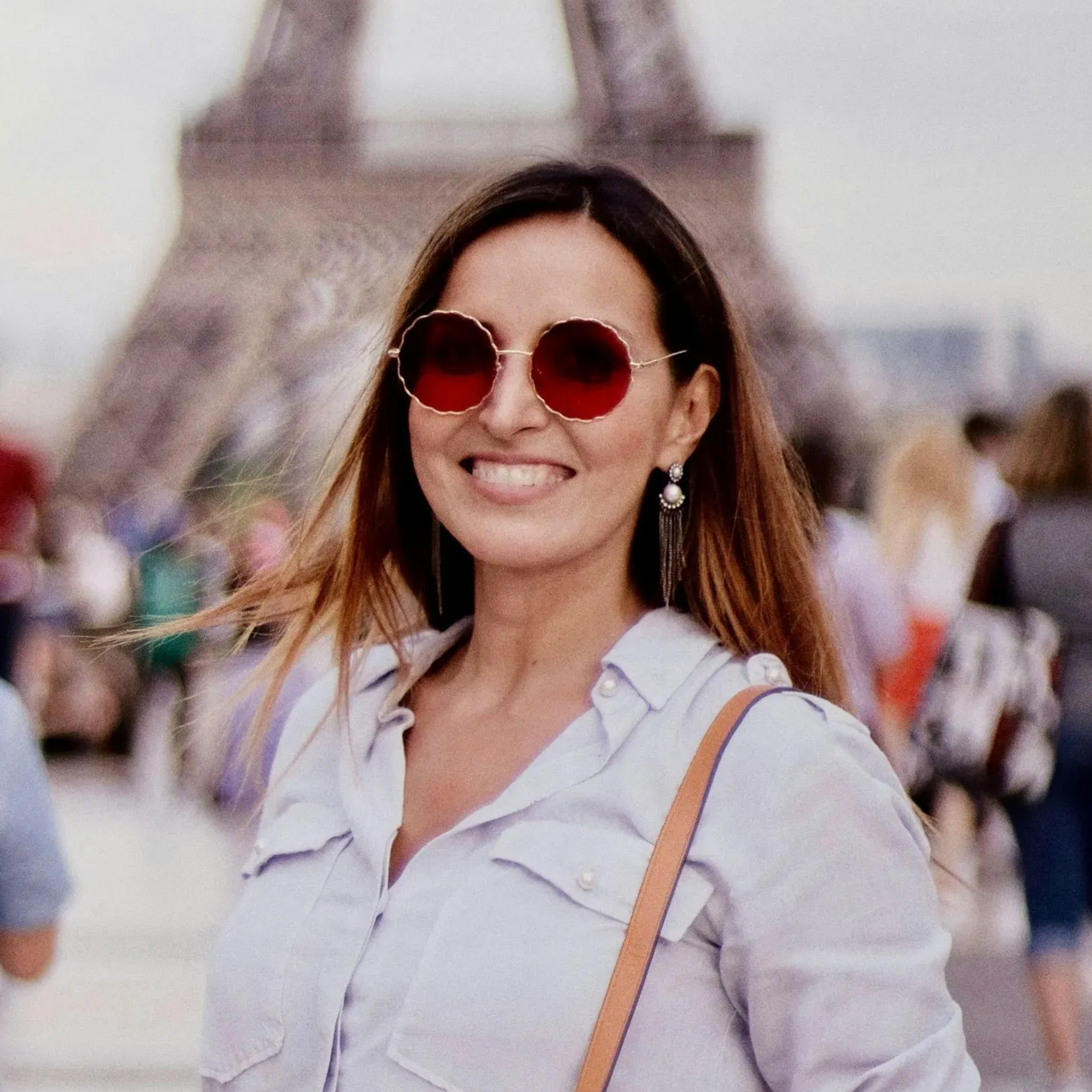 Woman with sunglasses smiles in front of the Eiffel Tower, wearing a light blue shirt.