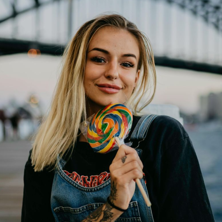 Woman with blonde hair holding a colorful lollipop, wearing overalls, and standing outdoors.