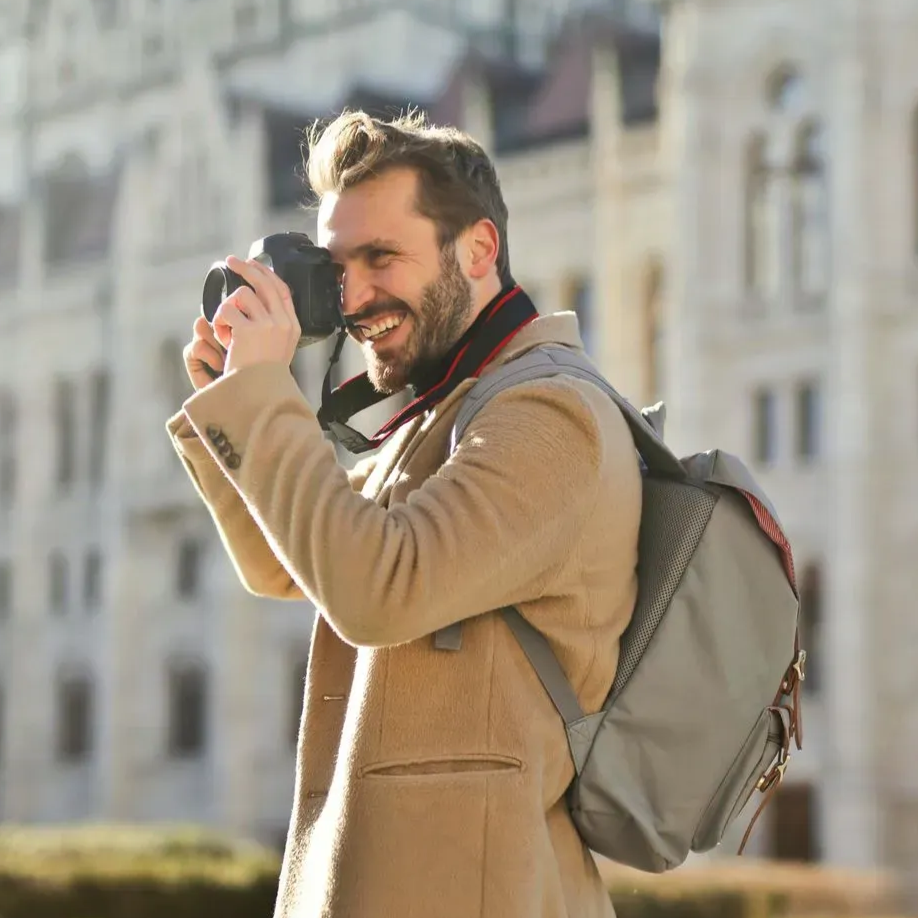 Man with camera smiles, backpack, tan coat, outdoors, taking photo of a building.