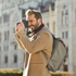 Man with camera smiles, backpack, tan coat, outdoors, taking photo of a building.