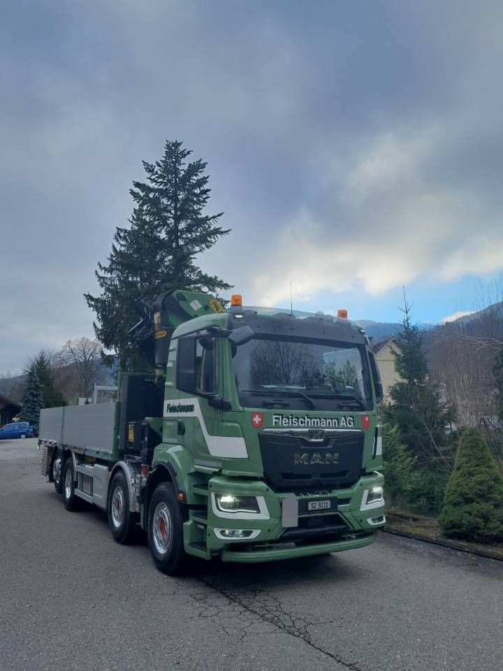 Grüner MAN-Lkw mit Kran auf einer Straße, bewölkter Himmel im Hintergrund.