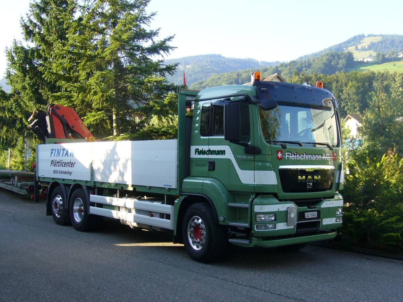 Grüner MAN-Lkw mit Kran auf einer Straße, vor der Kulisse von Bäumen und einem Hügelhang.