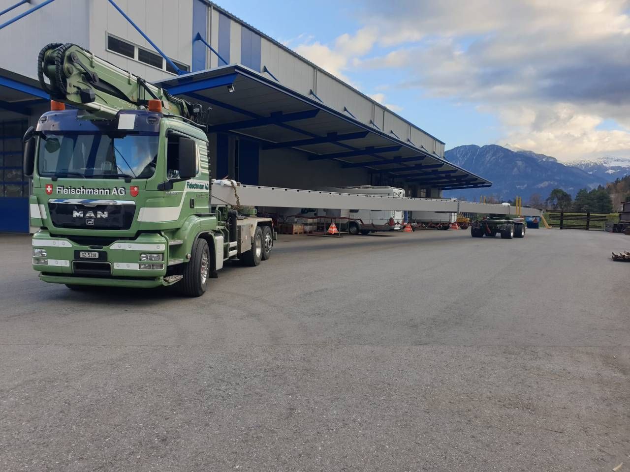Grüner LKW mit Kran transportiert eine lange Betonkonstruktion in der Nähe eines Lagerhauses; im Hintergrund sind Berge zu sehen.