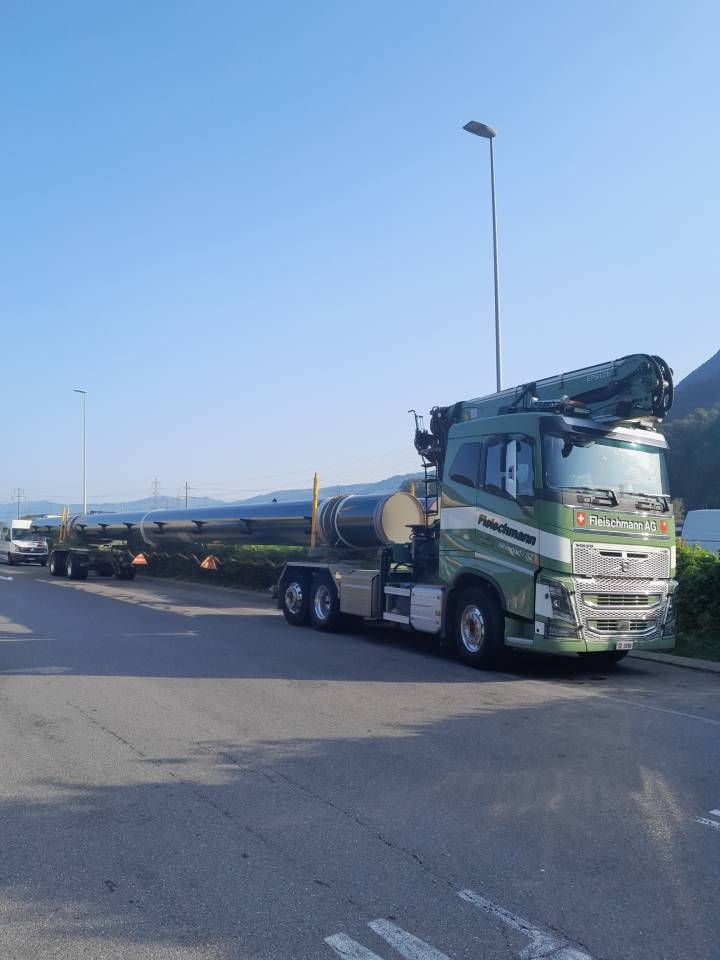 Green semi-truck hauling long black pipes, parked on asphalt; blue sky background.