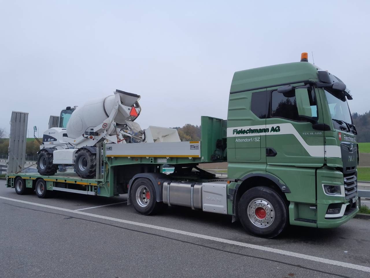 Ein grüner Sattelzug mit einem Betonmischer auf einem Tieflader steht am Straßenrand. Bewölkter Tag.