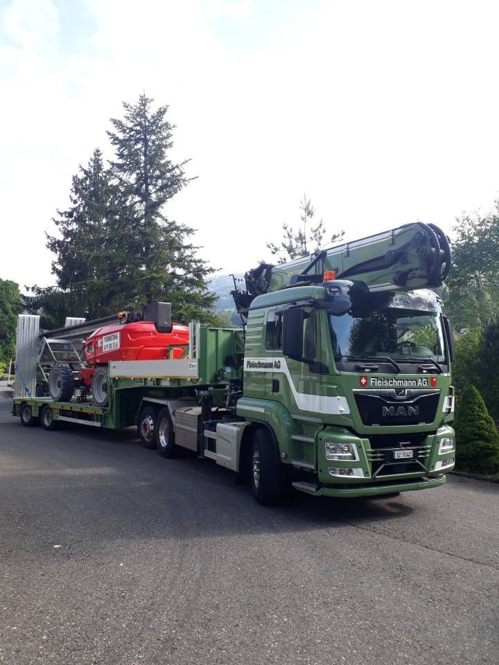 Ein grüner und weißer MAN-Lkw transportiert einen roten Traktor auf einem Tieflader. Im Freien, sonniger Tag.