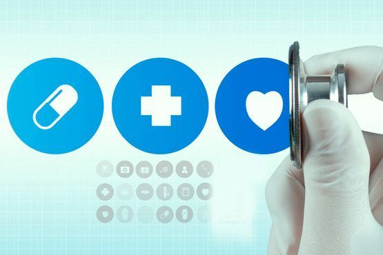 Una mano sostiene un estetoscopio cerca de iconos médicos azules: una pastilla, una cruz y un corazón.