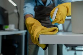 Persona con guantes amarillos y delantal limpiando un escritorio blanco con una botella de spray.
