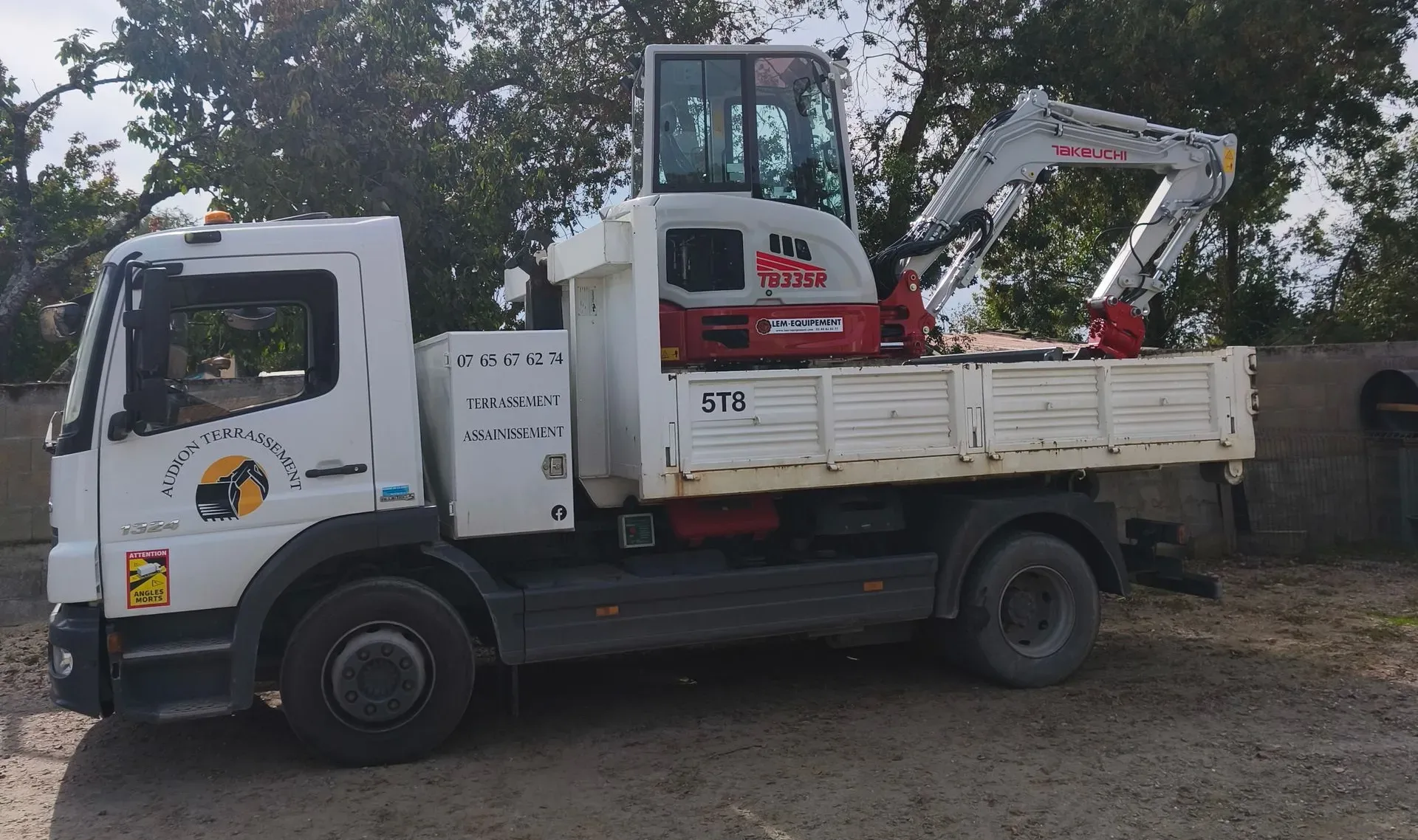 Camion plateau blanc avec une petite pelle mécanique rouge chargée sur sa benne. En extérieur, en journée.